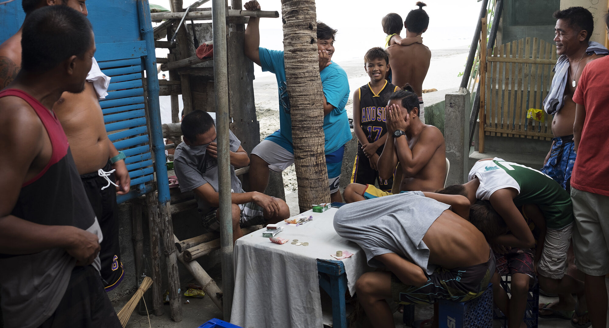 A mixed group of men and kids huddled around a table gambling in the late afternoon